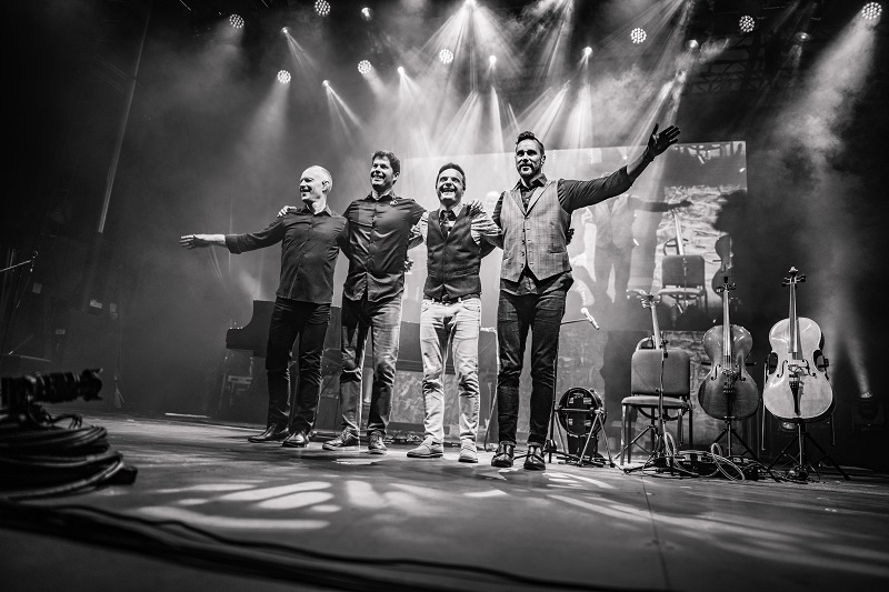 Black and white photo depicts four men bowing on stage with string instruments in background