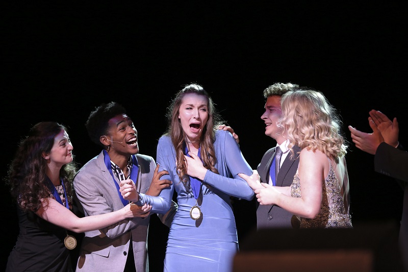 A young woman in a periwinkle dress cries tears of joy when she is surprised with an award on stage. Her peers surround her in celebration.