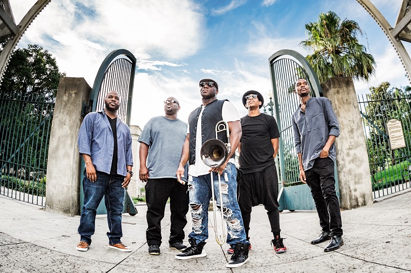 5 African American men stand together, one in the middle with a trombone, all wearing casual clothes like jeans and t-shirts. 