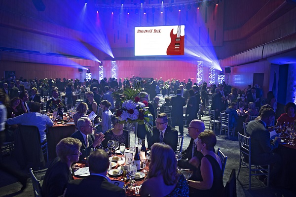 Hundreds of people sit at tables in Peter Kiewit Hall. The lights are blue and green with a shining projection of the Broadway Ball image on stage.