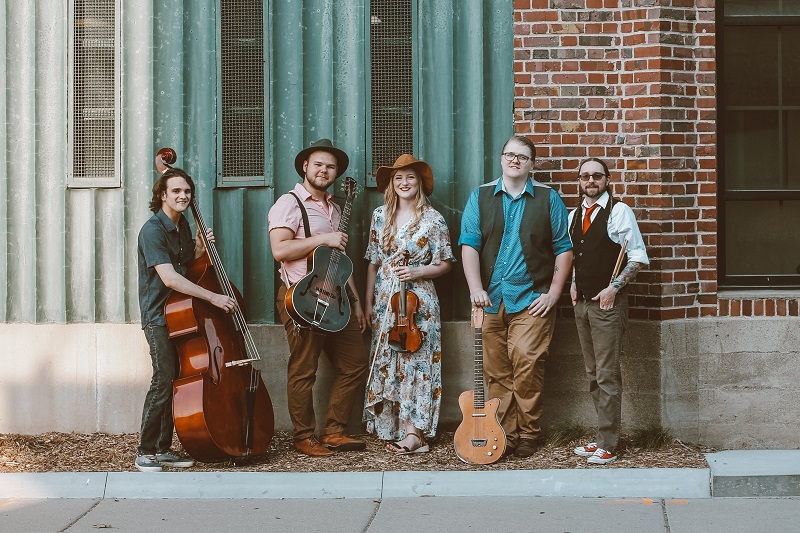 The Wildwoods band pose with their instruments in front of a steel building. Some members where cowbow hats. 