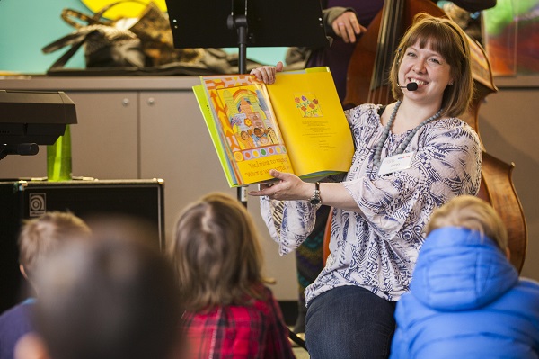 Kim Lomax sits on her knees in front of toddlers during a WeBop class. She's holding up a children's book and singing a jazz inspired song during story time.